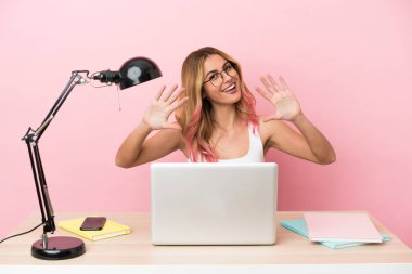 Pembe arka planda dizüstü bilgisayarı olan genç bir bayan öğrenci parmaklarıyla 10 tane sayıyor.