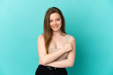 İzole edilmiş mavi arka planı olan genç bir kız bir ürünü sunmak için kenarı işaret ediyor.