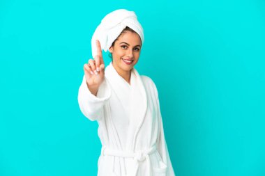 İzole edilmiş mavi arka planda bornozlu genç bir kadın parmağını kaldırıyor ve gösteriyor.