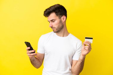 Genç beyaz yakışıklı adam sarı arka planda izole edilmiş cep telefonuyla kredi kartıyla alışveriş yapıyor.