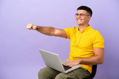 Genç adam dizüstü bilgisayarlı bir sandalyede oturuyor ve baş parmak hareketi yapıyor.