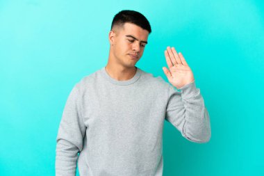 Soyutlanmış mavi arka planda genç yakışıklı adam dur hareketi yapıyor ve hayal kırıklığına uğruyor.