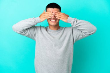 İzole edilmiş mavi arka planda genç yakışıklı adam. Gözleri elleriyle kaplıyor ve gülümsüyor.