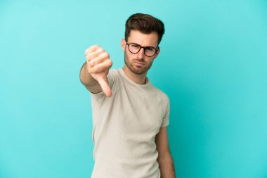 Mavi arka planda soyutlanmış beyaz, yakışıklı genç bir adam, olumsuz bir ifadeyle baş parmağını aşağı gösteriyor.