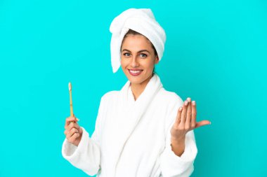 Bornozlu genç bir kadın, izole edilmiş mavi arka planda dişlerini fırçalıyor. Gelmene sevindim.