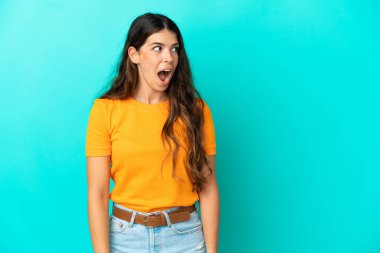 Mavi arka planda izole edilmiş genç beyaz kadın yan tarafa bakarken sürpriz jest yapıyor.