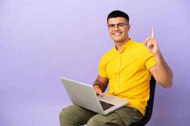 Genç adam dizüstü bilgisayarla bir sandalyede oturuyor ve en iyinin işareti olarak parmağını kaldırıyor.