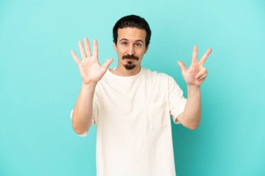 Mavi arka planda izole genç beyaz adam parmaklarıyla sekiz sayıyor
