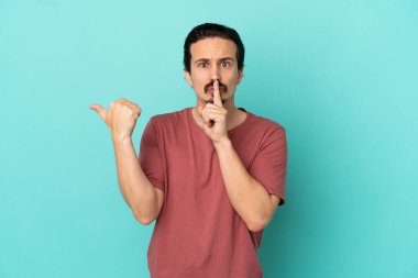 Kafkasyalı genç adam mavi arka planda izole edilmiş. Yan tarafı işaret ediyor ve sessizlik hareketi yapıyor.