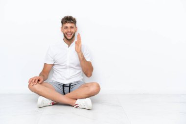 Genç sarışın adam beyaz arka planda izole bir şekilde yerde oturuyor ve ağzı açık bağırıyor.