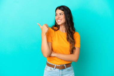 Mavi arka planda izole edilmiş genç beyaz kadın bir ürünü sunmak için kenarı işaret ediyor.