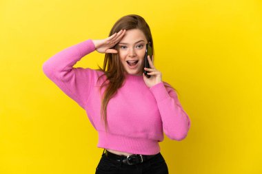 İzole edilmiş sarı arkaplan üzerinde cep telefonu kullanan genç kız sürpriz bir ifadeyle