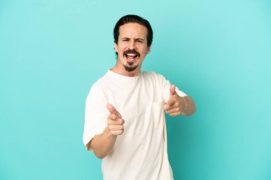 Mavi arka planda izole edilmiş genç beyaz adam ön tarafı işaret ediyor ve gülümsüyor.
