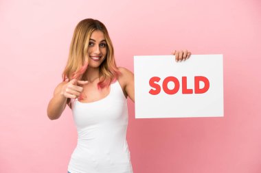 Soyutlanmış pembe arka planı olan genç bir kadın. Elinde yazılı bir pankart var ve ön tarafı işaret ediyor.