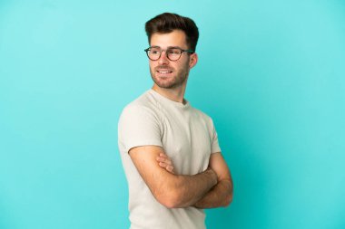 Mavi arka planda izole edilmiş, kolları bağlı ve mutlu, beyaz, yakışıklı genç bir adam.
