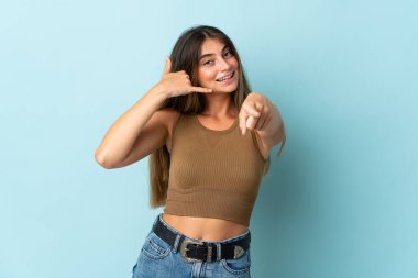 Mavi arka planda izole edilmiş genç beyaz kadın telefon hareketlerinde bulunuyor ve ön tarafı işaret ediyor.