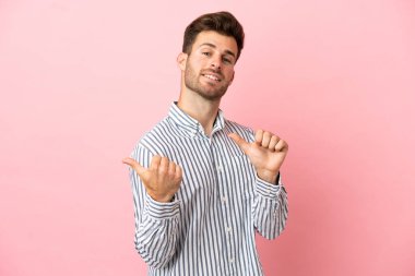 Pembe arka planda izole edilmiş beyaz, yakışıklı genç bir adam bir ürün sunmak için kenarı işaret ediyor.