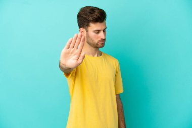 Beyaz tenli yakışıklı adam mavi arka planda izole edilmiş. Durma hareketi yapıyor ve hayal kırıklığına uğruyor.