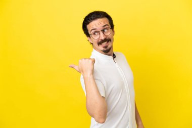 Sarı arka planda izole edilmiş genç beyaz bir adam bir ürün sunmak için kenarı işaret ediyor.