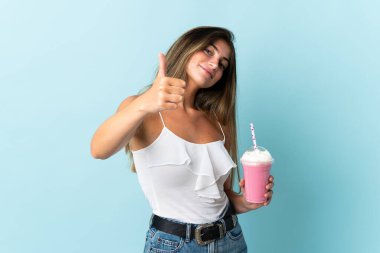 Çilekli milkshake 'i olan genç bir kadın mavi arka planda izole edilmiş, başparmakları havada çünkü iyi bir şey oldu.