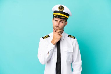 Kafkasyalı uçak pilotu mavi arka planda izole edilmiş. Şüpheleri var ve kafası karışmış bir yüz ifadesi var.