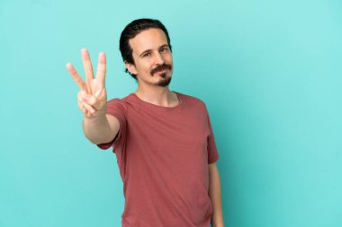 Mavi arka planda izole edilmiş genç beyaz adam mutlu ve parmaklarıyla üçe kadar sayıyor.