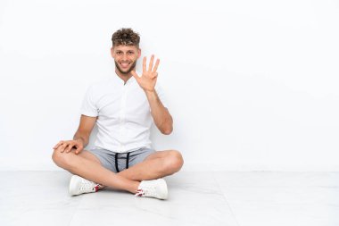 Genç sarışın adam beyaz arka planda izole bir şekilde yerde oturuyor mutlu ve parmakları olan dört kişi sayıyor.