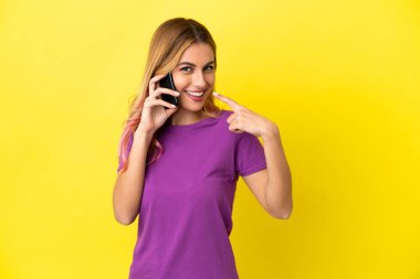 İzole edilmiş sarı arka planda cep telefonu kullanan genç bir kadın işaret veriyor.