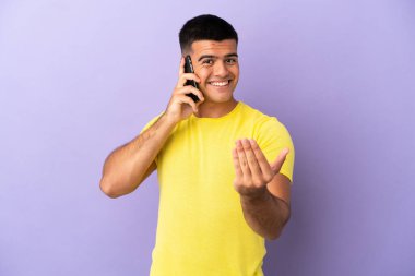 İzole edilmiş mor arka planda cep telefonu kullanan yakışıklı genç adam, davet edilmeye hazır. Gelmene sevindim.
