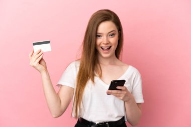 İzole edilmiş pembe arka planı olan genç bir kız cep telefonuyla alışveriş yapıyor ve elinde şaşırmış bir ifadeyle bir kredi kartı tutuyor.
