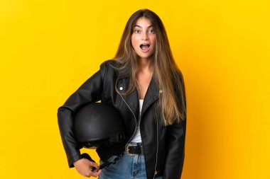Sarı arka planda, yüzünde sürpriz bir ifade olan motosiklet kaskı tutan kadın.