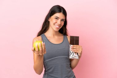 Pembe arka planda izole edilmiş beyaz bir kadın bir elinde çikolata tableti, diğerinde elma alıyor.