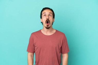 Kafkasyalı genç adam mavi arka planda izole edilmiş yukarı bakıyor ve şaşırmış bir ifadeyle