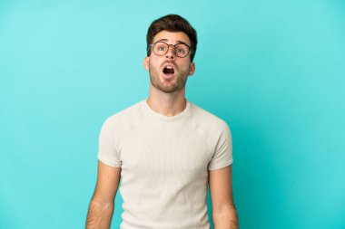 Kafkasyalı yakışıklı adam mavi arka planda izole edilmiş yukarı bakıyor ve şaşırmış bir ifadeyle