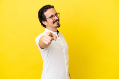 Sarı arka planda izole edilmiş genç beyaz adam mutlu bir ifadeyle ön tarafı işaret ediyor.