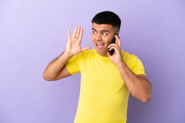 Mor arka planda cep telefonu kullanan genç yakışıklı bir adam kulağına elini sürerek bir şeyler dinliyor.