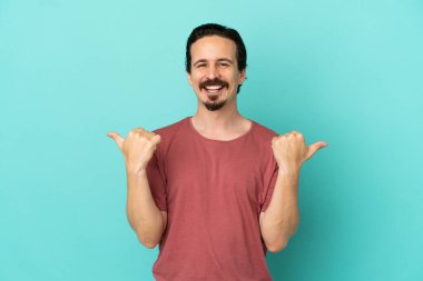 Kafkasyalı genç adam mavi arka planda izole edilmiş. Baş parmakları havada ve gülümsüyor.