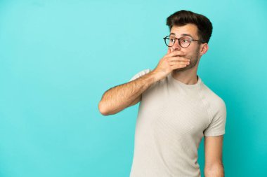 Genç, beyaz, yakışıklı bir adam mavi arka planda izole edilmiş. Yan tarafa bakarken sürpriz jest yapıyor.