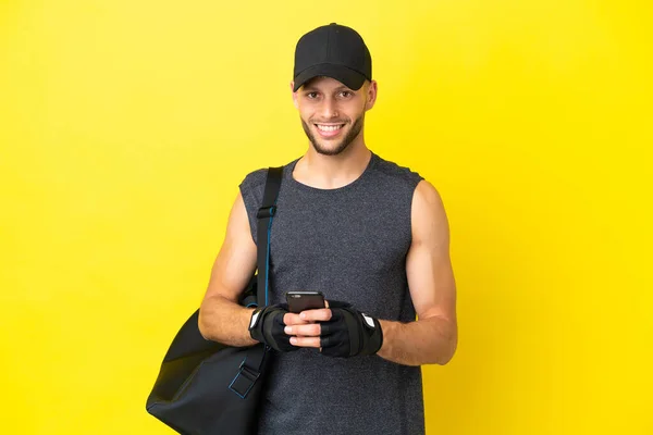 年轻的金发体育男子与体育袋隔离黄色背景发送消息与手机 — 图库照片