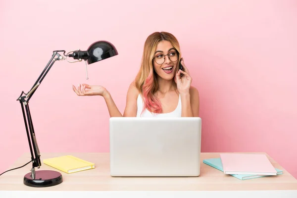 Young student woman in a workplace with a laptop over pink background keeping a conversation with the mobile phone with someone