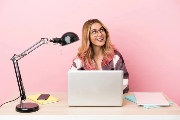Young student woman in a workplace with a laptop over pink background thinking an idea while looking up