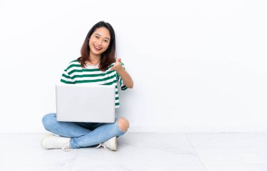 Yerde dizüstü bilgisayarı olan genç Vietnamlı bir kadın beyaz duvarda izole bir şekilde oturuyor ve iyi bir anlaşma için el sıkışıyor.