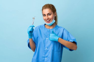 İzole edilmiş mavi arka plan üzerinde aletleri tutan genç bir dişçi, baş parmak işareti yapıyor.