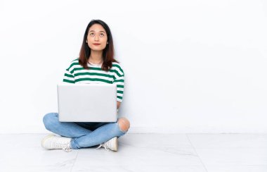 Diz üstü bilgisayarı olan genç Vietnamlı kadın beyaz duvarda izole bir şekilde oturuyor ve yukarı bakıyor.