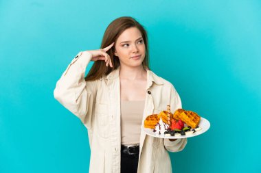 İzole edilmiş mavi arka planda waffle tutan genç kız kuşkuları ve düşünceleri var.