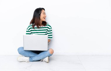 Yerde dizüstü bilgisayarı olan genç Vietnamlı bir kadın beyaz duvarda izole bir şekilde oturuyor ve yan pozisyonda gülüyor.