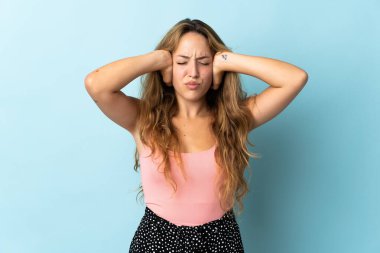 Mavi arka planda izole edilmiş genç sarışın kadın hayal kırıklığına uğramış ve kulaklarını kapatmış.