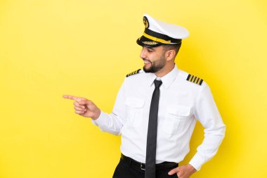 Uçak Arap pilotu, sarı arka planda izole edilmiş, parmağını yana doğru gösteren ve bir ürün sunan.