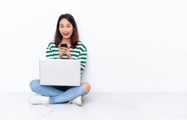 Yerde dizüstü bilgisayarı olan genç Vietnamlı bir kadın beyaz duvarda izole edilmiş, şaşırmış ve bir mesaj gönderiyor.