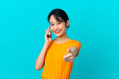 Genç Vietnamlı kadın mavi arka planda izole edilmiş. Cep telefonuyla konuşurken baş parmağını kaldırıyor.